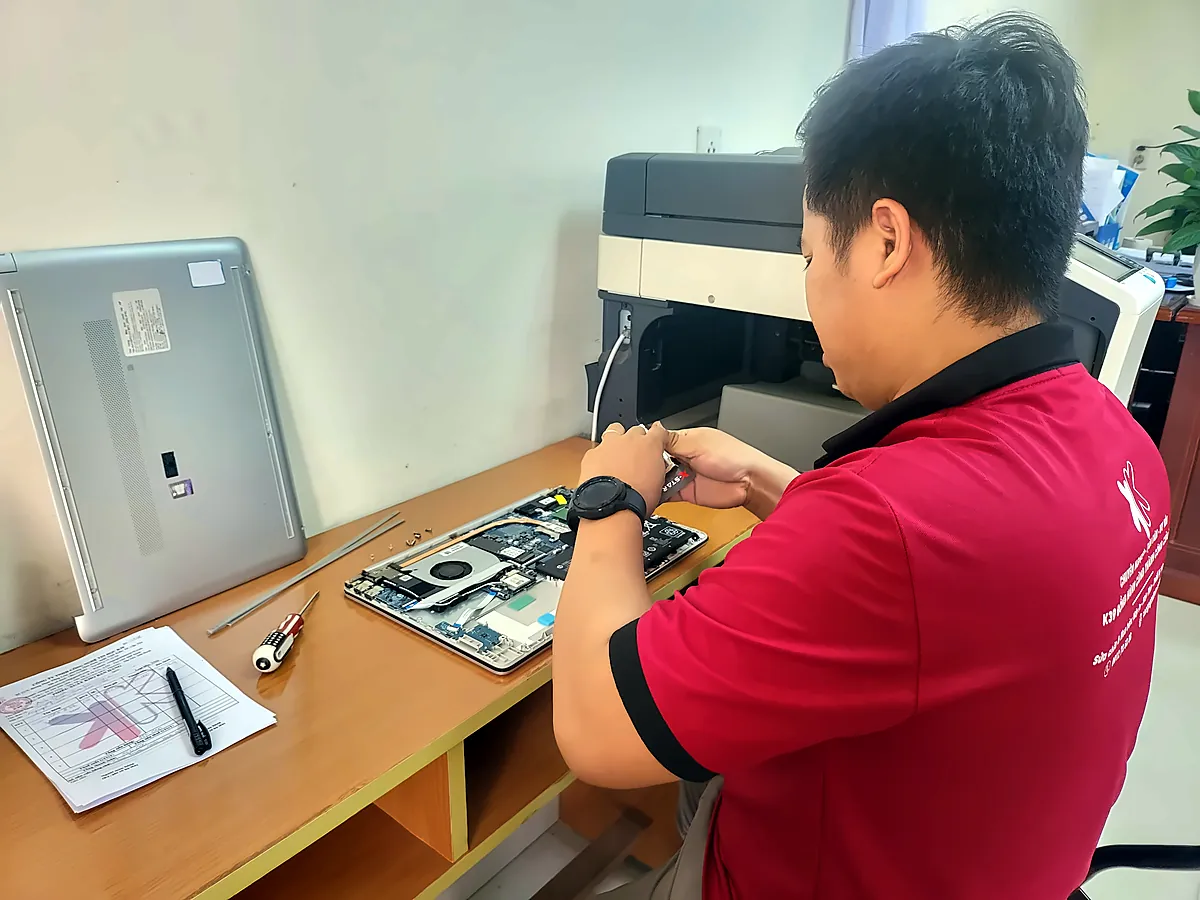 Kỹ thuật viên K39 kiểm tra, sửa laptop tại bàn làm việc, bên cạnh máy in